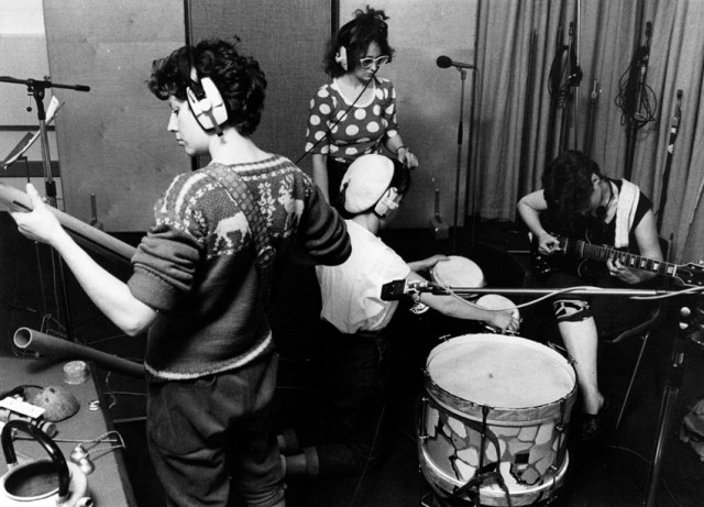 Four members of Cast Iron Fairies in recording studio, playing guitars and bongo drums and wearing earphones. Microphones and a large bass drum also shown.