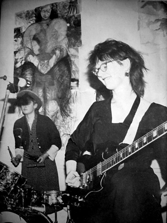 Black and white photo of two of the Cast Iron Fairies, Babs on left playing a drum kit and hand-held percussion instruments and standing behind a microphone, Jackie playing guitar and smiling. A large batik backdrop wall-hanging made by the band shows a large figure of a seated woman.