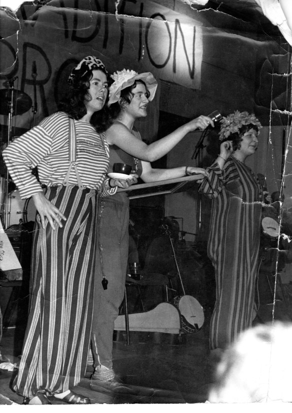 The three band members stand on stage in the middle of a performance. They wear wacky costumes. Behind them are a stage of instruments and banners (writing not decipherable)