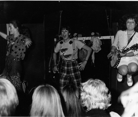 Three members of the band perform in front of a crowd. Black and white. The image includes the back of the audience's heads. Rae is jumping in the air and plays the banjo. They wear vibrant costumes.