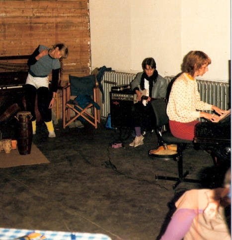 Val  dancing expressively, Sally on guitar and Maggie playing piano, with microphones, amplifiers and costumes also in photo.