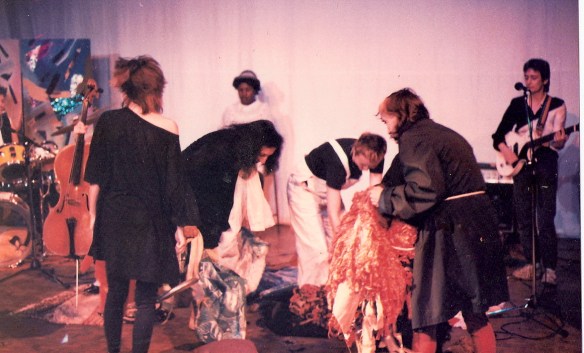 Contradictions on stage, some women picking up brightly coloured costumes, others playing guitar and drums, other instruments also in photo include cello. Handpainted screens in background.