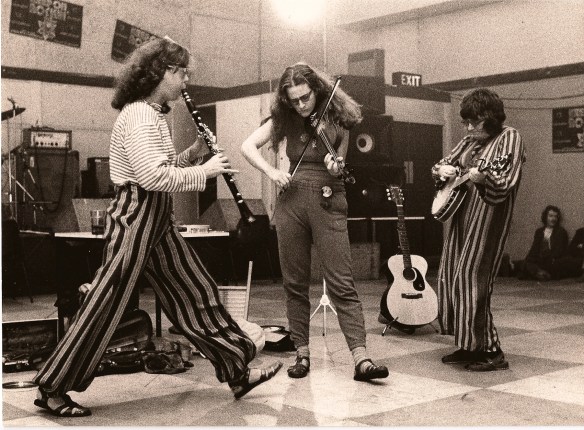 Another picture depicting musical performance. Caroline plays a clarinet, Rix a violin and Rae a banjo. The venue looks like a community centre.