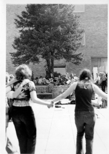 Women dancing in a huge outdoor circle, Susan with guitar in background under a tree, women are smiling and laughing, holding hands.