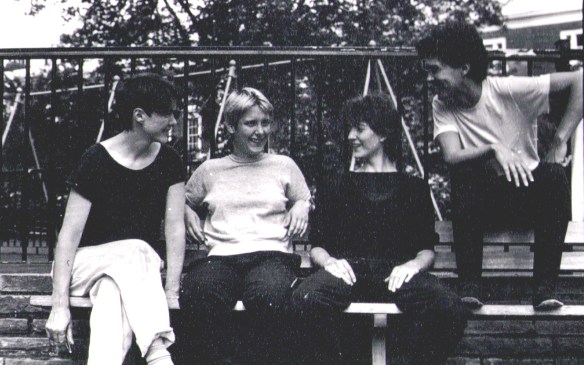 The four women in a black and white photo in a children's playground, smiling and seated on a bench, talking to one another.
