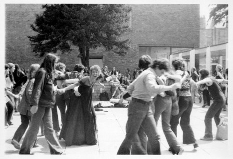Women dancing in  huge circle as Susan sings and plays guitar. Women are holding hands or have their arms around one another's shoulders, laughing and smiling.