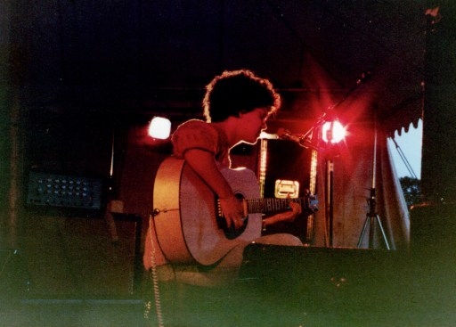 A colour photo of Georgette Okey on stage, playing her acoustic guitar in a marquee, with bright red lighting, daytime sky just visible through the tent door in the background..
