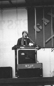 Carole on stage with her keyboards and singing, and huge Gay Pride banners behind.