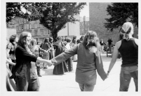 Women dancing in  huge circle as Susan sings and plays guitar. Women are holding hands or have their arms around one another's shoulders, laughing and smiling.