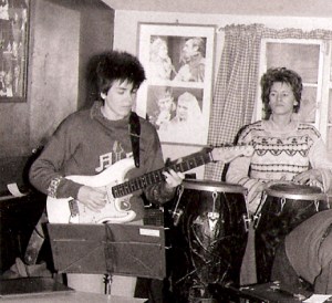 Deirdre playing guitar at a Guest Stars rehearsal, looking seriously at her fret board while Linda playing congas watches her. 