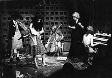 Black and white photo of five Feminist Improvising Group members, playing trumpet, double bass, keyboards and singing, a performance in action.