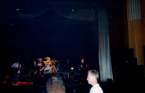 A live gig on stage with spotlights on the band and audience dancing in front.