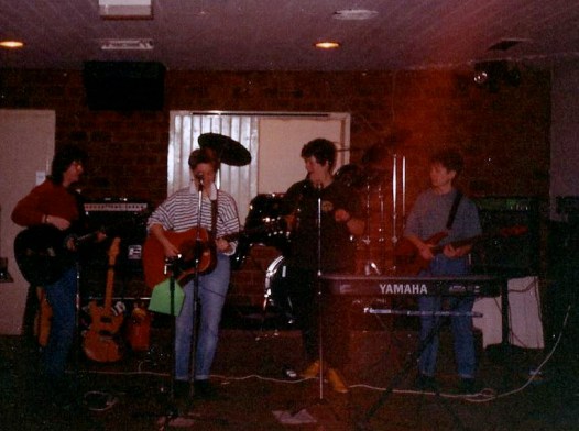 The four Fabulous Jam Tarts in concert, a darkened stage, guitarists and singer at front.
