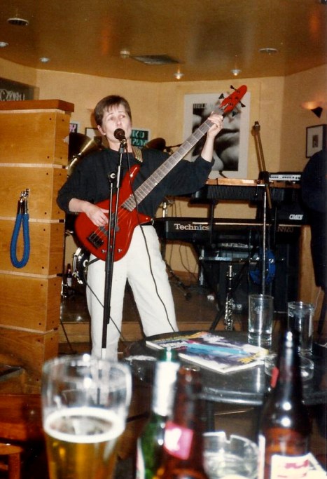 bass player on stage, playing and singing, pints of beer on table in front of stage indicate a pub gig.