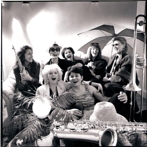 A black and white publicity shot of Hi Jinx, posing in a tropical setting. The seven women are sitting close together on a sofa, some with arms around each other, laughing and grinning, under bright lights, with a huge beach umbrella, straw hats, palm tree branches, saxophones and a trombone.