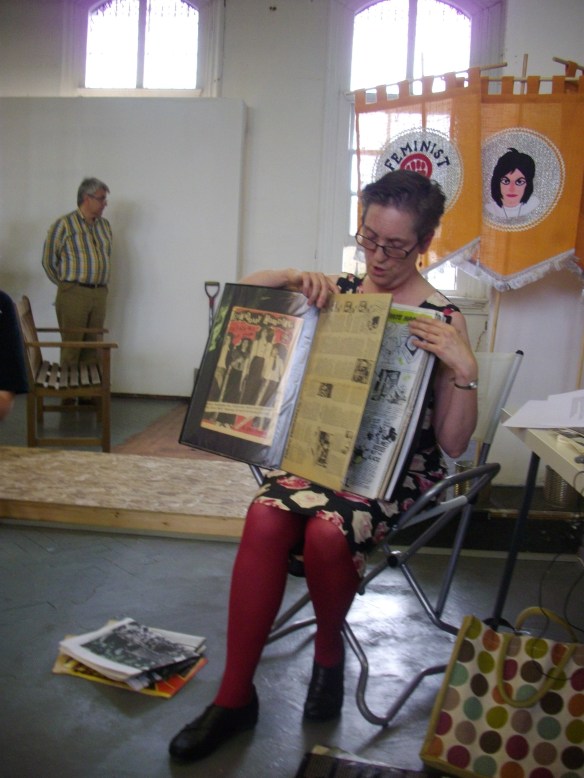 Lucy Whitman shares the contents of her scrapbook which includes the publications Jolt and Drastic Measures. She holds the pages of the scrapbook open and talks about them. In the background a visitor to the exhibition, and a WLM symbol on the Feminist Disco banner.
