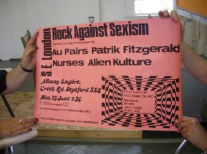 Poster from Lucy Whitman's collection of Rock Against Sexism memorabilia held up and photographed during a presentation. Pink background, black writing says 'Rock Against Sexism, South East London, benefit for Gay Pride 1980. The Au Pairs, Patrik Fitzgerald, Nurses, Alien culture. At the Albany Empire, SE8. 23 June. £2/1.50.'