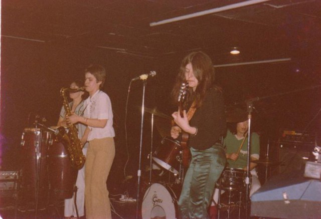 Isabel on conga drums, Angele playing sax, Laka on guitar and vocals, with Sheila behind them on drum kit, performing on a dimly lit stage.