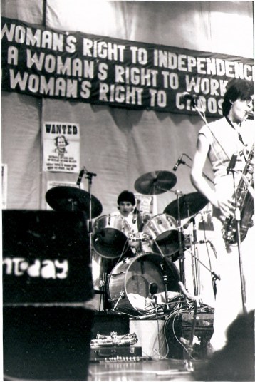 Jackie, Jam Today's drummer, and Julia, saxophonist, playing intently. A large banner above the stage reads 'A WOMAN'S RIGHT TO INDEPENDENCE, A WOMAN'S RIGHT TO WORK, A WOMAN'S RIGHT TO CHOOSE.' Behind the drum kit is a 'WANTED' poster bearing Thatcher's face.