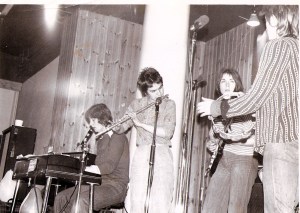 Four members of Jam Today on stage, looking up from the audience's point of view. Keyboard, flute and guitar players and the singer playing tambourine looking at the guitarist. Amplifiers in background, microphones in front.