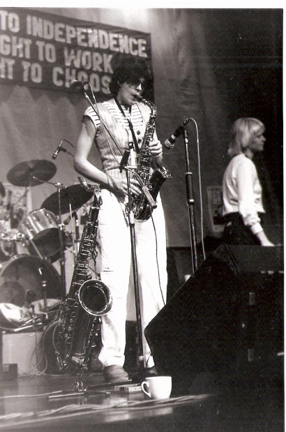 Julia is playing the saxophone and Barbara singing, into microphones at the front of the stage, and Jackie's drum kit is visible behind them. A large banner above the stage reads 'A WOMAN'S RIGHT TO INDEPENDENCE, A WOMAN'S RIGHT TO WORK, A WOMAN'S RIGHT TO CHOOSE.'