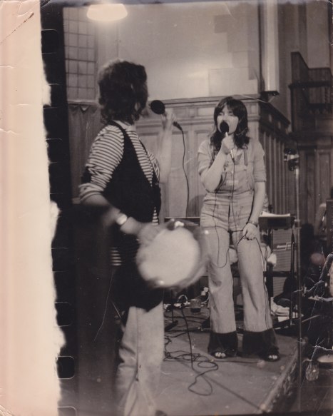 A faded sepia photo of Luchia and Angela on a small, curtain-draped stage at the social of the Edinburgh national WLM conference. The two singers are each holding a microphone and singing while looking at each other intently, and Luchia also plays a tambourine.