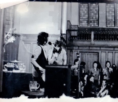 A second photo of Luchia and Angela on stage, singing, with women audience members watching from the side of the hall.