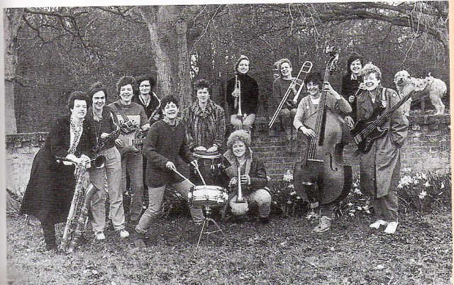 In a black and white publicity shot, thirteen women from the band, and a dog, stand outside in a walled garden with their instruments, laughing.