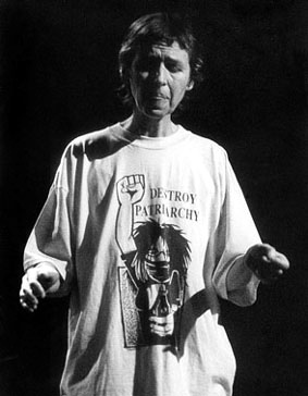 Black and white photograph of Maggie Nicols singing, focusing with eyes closed and hands outstretched expressively, wearing a t-shirt with a cartoon of a spiky-haired girl raising a large clenched fist and the words 'Destroy Patriarchy.'
