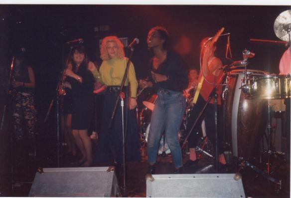 A dynamic picture of several women of Meet Your Feet on stage in performance, singing and playing electric bass guitar and guitar.