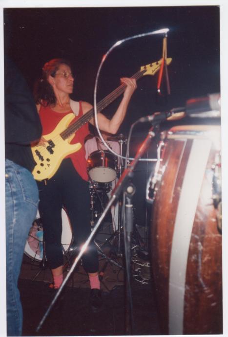 Bass guitar player in performance, conga drums, drum kit and mikes on stage.