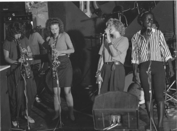 Photo of the band performing in a pub showing sax and clarinet players, singers and drum kit, amplifiers and microphones.