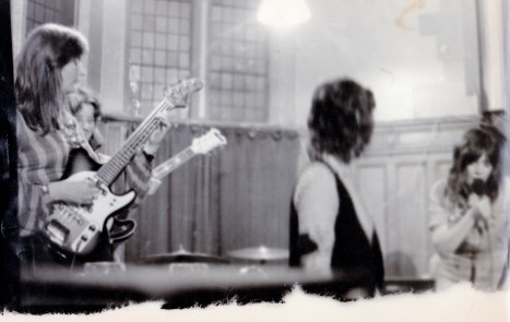 A tattered close up photo of the NWLRB performing, Angela singing intently into microphone, watched by Luchia, and Jenny and Frances playing behind them.