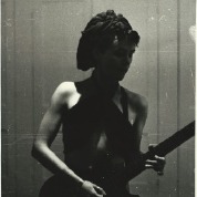 Pauline Channing playing guitar, wearing black teeshirt and cap, silhouetted.