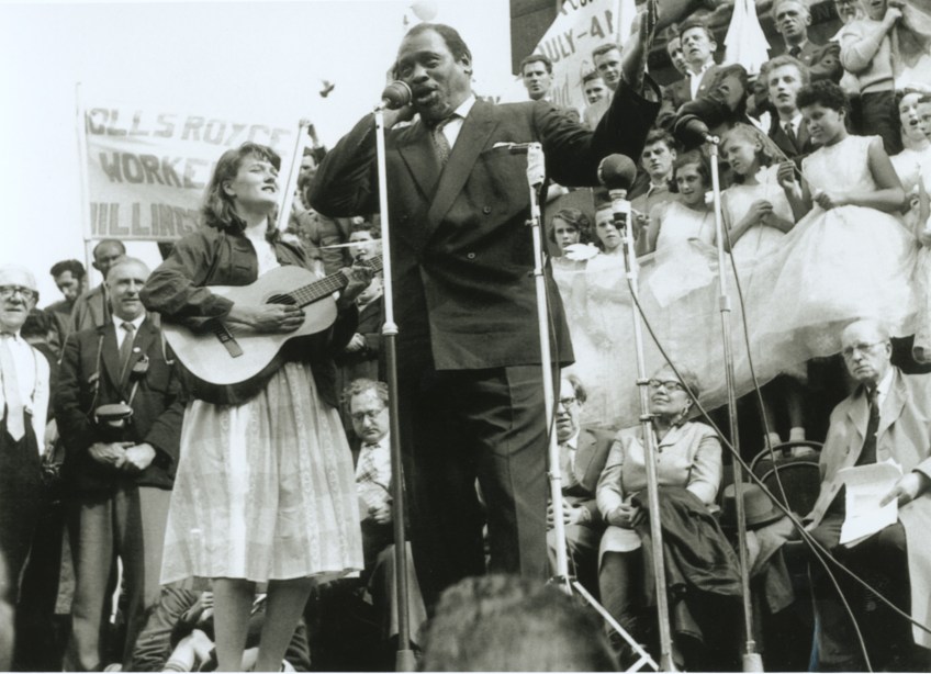 Peggy on the plinth in Trafalgar Square in 1960, playing the guitar next to Paul Robeson who is singing, one hand over his ear. Crowds and placards in background, but protest unknown.