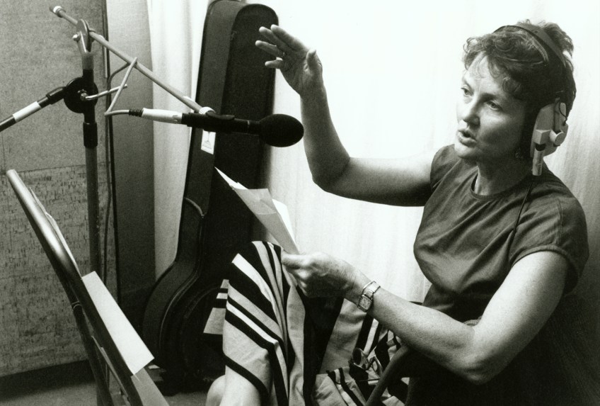 Peggy in recording studio, singing into a micrphone, guitar case in background and music sheet on her knee.