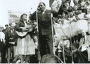 Peggy plays guitar and sings on stage with Paul Robeson in Trafalgar Square. Placards and crowds in the background but not clear what the demonstration is.