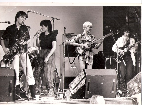PMT in performance, saxophonist, singer holding microphone and looking at the drummer at back of stage, bass guitarist smiling, guitarist looking seriously at her guitar. All have microphones and there are a lot of amplifiers on stage.