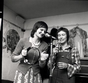 Two women in rehearsal. Ruthie plays the tambourine and uses a microphone on a stand, and Caroline sings into a hand held mike. Both are clearly enjoying themselves.