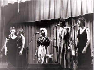 Five women singers on stage with microphones and amplifiers, dressed in dresses and smart trousers.