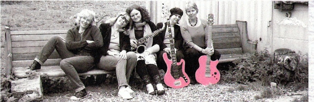 The five Stepney Sisters in 2011, all seated on a long wooden bench, with their instruments, leaning against one another affectionately and smiling. The bass and guitar are picked out in bright pink.