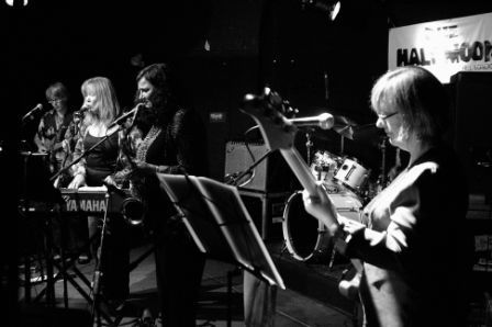 A photo of the reunited band, on stage playing guitar, singing, keyboards, and bass, the drum kit at the back of the stage and music stands, microphones and amplifiers also.
