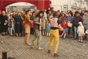 Colour action photo of the band playing on the street. An audience watches as they play. 