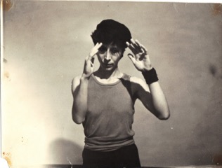 Nina in a black and white photo, wearing vest and black cap, both hands raised in a salute-like pose.