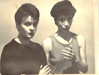 Black and white photo: pauline Channing in black t-shirt, Nina Rapi in vest and cap, looking serious, holding hands in front of their chests.