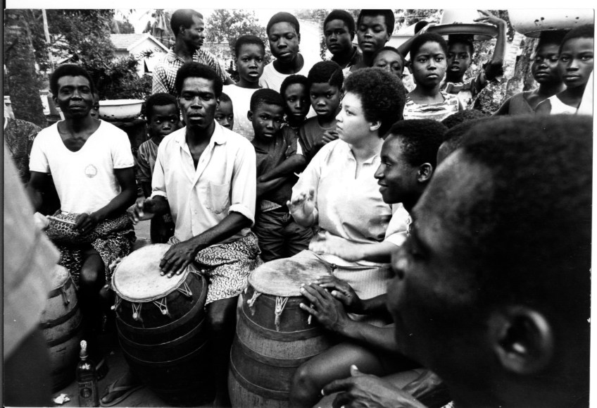 Terri Quaye playing conga drums with several other drummers seated in a circle and an audience standing in background.