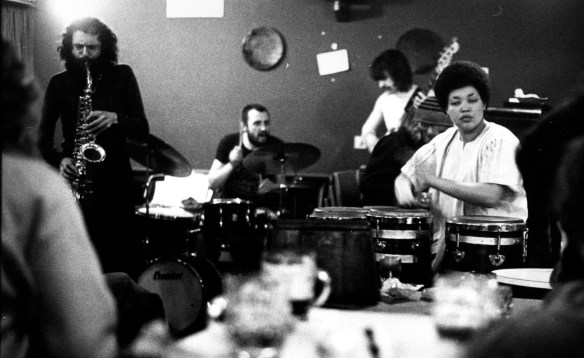 Terri Quaye playing conga drums on stage with three musicians on sax, drums and guitar.