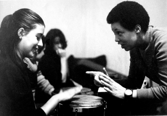 Terri Quaye and a pupil in profile with hands over a conga drum, other pupils in background.