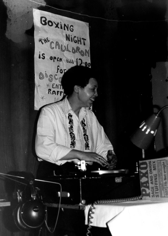 Terri Quaye in DJ mode at her Cauldron Women's Disco, working turntables, a hand-painted poster advertising the Boxing Night event and raffle on the wall behind. 