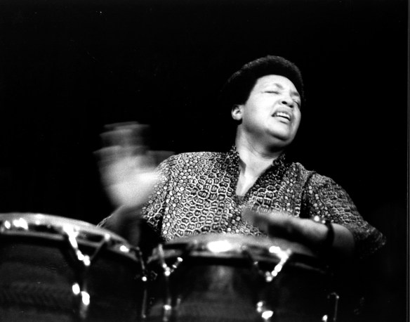 Terri Quaye playing a solo concert on conga drums, hands blurred with speed. 